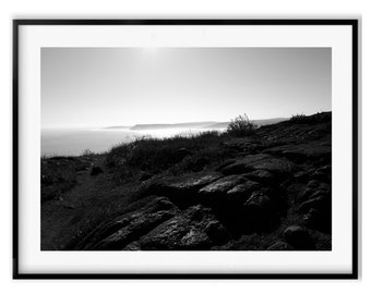 May include: A black and white photograph of a rocky coastline with the ocean in the distance. The sun is shining brightly in the sky, creating a hazy effect over the water.