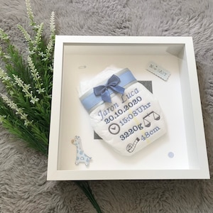 Könnte beinhalten: Ein weißer Bilderrahmen mit einer bestickten Windel. Die Windel ist mit einem blauen Band verziert und zeigt den Namen "Jaron Luca", Geburtsdatum, Uhrzeit, Gewicht (3220g) und Größe (48cm). Ein Giraffenaufkleber ist ebenfalls enthalten.