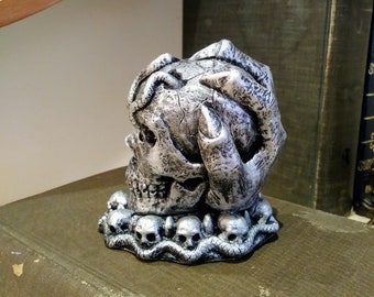 May include: A silver skull sculpture with a hand holding its head. The skull is surrounded by a ring of smaller skulls.