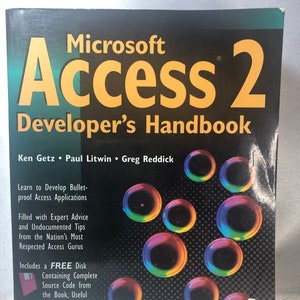 Op de afbeelding: Een zwart boek met een gele titel die "Microsoft Access 2 Developer's Handbook" luidt. De omslag van het boek heeft een kleurrijk ontwerp van overlappende cirkels op een zwarte achtergrond.