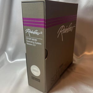 May include: A taupe-colored rectangular software box for the Rainbow CP/M-86/80 Operating System Version 2.0. The box features purple horizontal stripes, the Rainbow logo in white script, and a circular sticker labeled Classified Software.