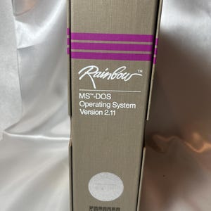 May include: A rectangular beige box for the Rainbow MS-DOS Operating System Version 2.11. The box features two horizontal purple stripes, the Rainbow logo, the text MS-DOS Operating System Version 2.11, a circular silver seal, and the word digital.