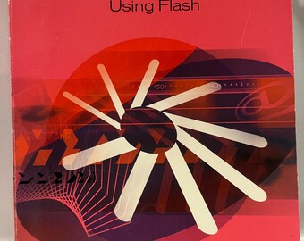 May include: A book cover for Macromedia Flash 5, with the title "Using Flash." The cover is primarily red with a large white and purple graphic design. The Macromedia logo is at the bottom right.