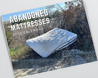 May include: A calendar titled "ABANDONED MATTRESSES 2026 CALENDAR" features a white mattress with black trim, resting outdoors. The image includes text at the bottom: "CREATED BY SHUTUPANDTAKEMYMONEY.COM". The calendar is set against a backdrop of foliage and a blue sky.