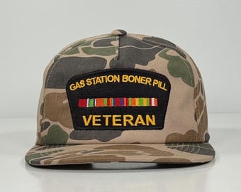 May include: A camouflage print baseball cap with a flat brim. The cap features a patch with the text "GAS STATION BONER PILL" above a row of colorful stripes and the word "VETERAN" below.