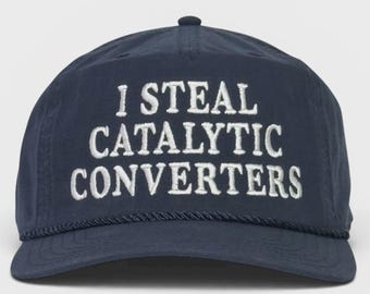 May include: A navy blue baseball cap featuring the white embroidered text I STEAL CATALYTIC CONVERTERS across the front panel. The cap includes a matching navy blue braided cord detail along the base of the crown and a curved brim.