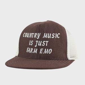 May include: A brown trucker hat with a white mesh back and a curved brim. The hat features the embroidered text "COUNTRY MUSIC IS JUST FARM EMO" in white, centered on the front panel. The hat is a casual accessory.