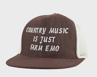 May include: A brown trucker hat with a white mesh back and a curved brim. The hat features the embroidered text "COUNTRY MUSIC IS JUST FARM EMO" in white, centered on the front panel. The hat is a casual accessory.