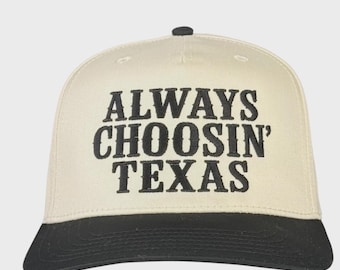 May include: A cream-colored baseball cap with a black brim and the embroidered text "ALWAYS CHOOSIN' TEXAS" in bold, black letters. The cap has a classic trucker hat design.