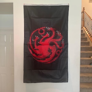 May include: A rectangular black wall flag featuring a large, centred red graphic of a three-headed dragon coiled in a circular shape. The flag has a white reinforced header strip along the top edge with two metal grommets for hanging purposes.