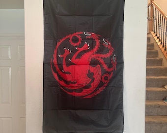 May include: A rectangular black wall flag featuring a large, centered red graphic of a three-headed dragon coiled in a circular shape. The flag has a white reinforced header strip along the top edge with two metal grommets for hanging purposes.
