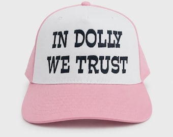 May include: A baseball cap featuring a white front panel with the black embroidered text IN DOLLY WE TRUST. The cap has a light pink brim and side panels, a structured crown, and a button on top. The design is centered on the front panel.