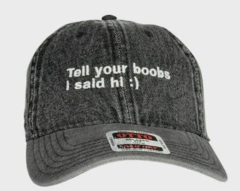May include: A black denim baseball cap with the text "Tell your boobs I said hi :)" embroidered in white. The cap has a curved brim and an adjustable strap.