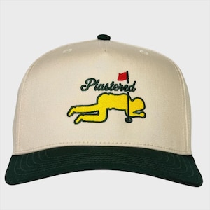 Puede incluir: Gorra de béisbol beige con visera verde oscuro y botón verde. La gorra presenta la palabra "Plastered" en escritura verde sobre una figura amarilla tumbada junto a una bandera de golf.