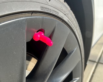 May include: A close-up of a black car tire with a pink valve stem cap. The tire has the text "FLIES/SIDEWALL POLYESTER 2 + STEEL 2 + NYLON 1 TREAD POLYESTER 2 + STEEL 2 + NYLON 1" printed on the sidewall.
