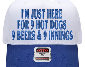 May include: A white and blue baseball cap with the text "I'M JUST HERE FOR 9 HOT DOGS 9 BEERS & 9 INNINGS" embroidered in blue. The cap has a curved brim and a button on top. The brand tag is visible on the brim.