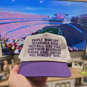 Pode incluir: Boné de caminhoneiro branco e roxo com o texto: "PEOPLE WHO GET DIARRHEA FROM TACO BELL ARE WEAK AND THEIR BLOODLINE IS WEAK AND HISTORY WILL FORGET THEM." O boné tem uma coroa branca, uma aba roxa e um detalhe de corda branca.