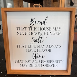 May include: A wooden framed sign with a white background and black text. The text reads "Bread that this house may never know hunger. Salt that life may always have flavor. Wine that joy and prosperity may reign forever."