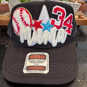 May include: Black trucker hat with a baseball, stars, and the number 34. The word "Mama" is embroidered in white script. The hat features a foam front and a mesh back.