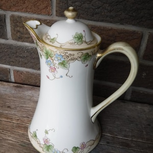 Puede incluir: Una cafetera de porcelana blanca con ribete dorado y diseño floral. La cafetera tiene una tapa con un pomo en la parte superior. La cafetera está decorada con flores y hojas rosas, azules y verdes.