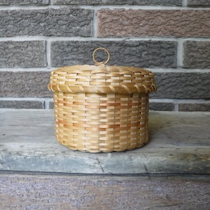 Può includere: Un piccolo cestino rotondo intrecciato con coperchio. Il cestino è realizzato in vimini marrone chiaro e ha una maniglia circolare sulla parte superiore. Il cestino è appoggiato su una superficie di legno stagionato davanti a un muro di mattoni.