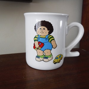 May include: White ceramic mug featuring a cartoon illustration of a Cabbage Patch Kid holding a red toy car. The mug has a white handle and a yellow toy car on the side.