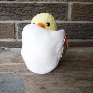 May include: A yellow crocheted chick peeks from a white plush egg. The chick has black eyes and an orange beak. The egg is open, and the chick is nestled inside. The background is a brick wall and a wooden surface.