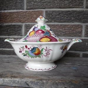 Puede incluir: Un plato de servir de porcelana blanca con tapa, que presenta un patrón floral colorido con frutas y una cesta. El plato tiene un borde festoneado y una base decorativa.