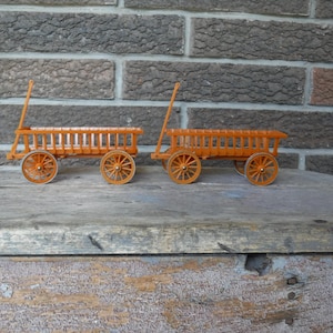 May include: Two miniature orange wooden wagons with spoked wheels and a handle. The wagons are displayed on a weathered wooden surface against a brick wall background. The wagons are likely decorative items.