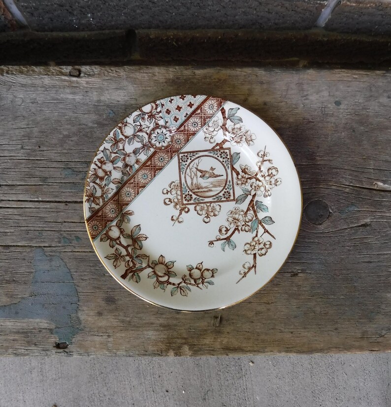 Puede incluir: Un plato redondo de cer&aacute;mica con base color crema que presenta un patr&oacute;n de ramas florales en marr&oacute;n y azul verdoso. Una banda diagonal con motivos geom&eacute;tricos cruza la parte superior izquierda y un marco cuadrado central muestra un ave.