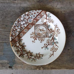 Puede incluir: Un plato redondo de cer&aacute;mica con base color crema y un patr&oacute;n floral en marr&oacute;n y azul claro. El dise&ntilde;o incluye un marco cuadrado central con la ilustraci&oacute;n de un p&aacute;jaro, rodeado de ramas florecidas y un borde geom&eacute;trico.