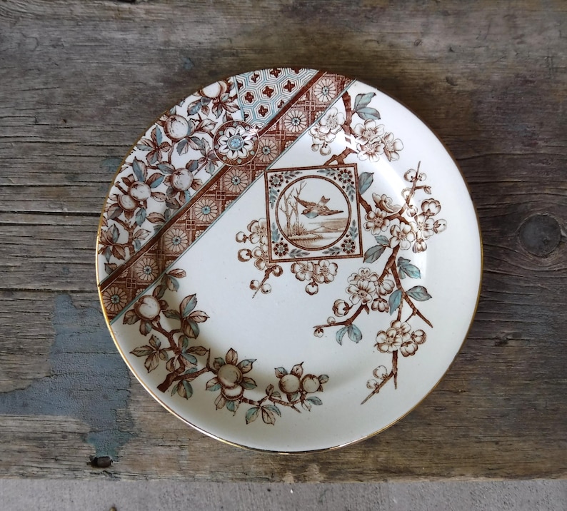 Puede incluir: Un plato redondo de cer&aacute;mica con base color crema y un patr&oacute;n floral en marr&oacute;n y azul claro. El dise&ntilde;o incluye un marco cuadrado central con la ilustraci&oacute;n de un p&aacute;jaro, rodeado de ramas florecidas y un borde geom&eacute;trico.
