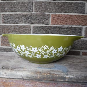 May include: A vintage green mixing bowl with a white floral pattern around the exterior. The bowl has a wide rim and a white interior. It is sitting on a weathered wooden surface in front of a brick wall.
