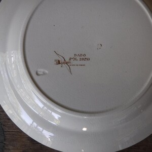 Puede incluir: La parte inferior de un plato de cer&aacute;mica blanca que presenta un sello de color marr&oacute;n oscuro con el texto DADO y Rd No. 20250, junto con una peque&ntilde;a marca impresa. La superficie es lisa con un acabado vidriado sutil y un borde ancho.