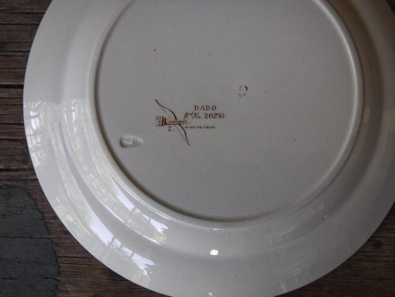 Puede incluir: La parte inferior de un plato de cer&aacute;mica blanca que presenta un sello de color marr&oacute;n oscuro con el texto DADO y Rd No. 20250, junto con una peque&ntilde;a marca impresa. La superficie es lisa con un acabado vidriado sutil y un borde ancho.