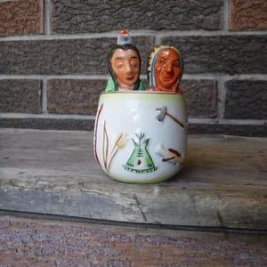 Puede incluir: Una taza de cerámica vintage con dos figuras nativas americanas. La taza blanca presenta detalles pintados, incluyendo un tipi, un tomahawk y motivos florales. Las figuras tienen rasgos faciales y tocados detallados.