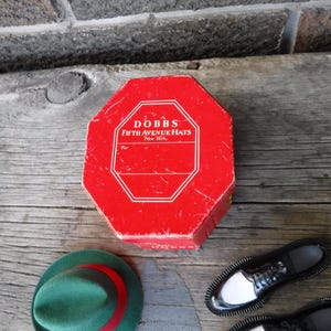 Op de afbeelding: Een achthoekige rode hoedendoos met de tekst "DOBBS Fifth Avenue Hats New York" staat op een houten oppervlak. Een groene hoed met een rode band en zwart-witte schoenen suggereren een vintage modethema.