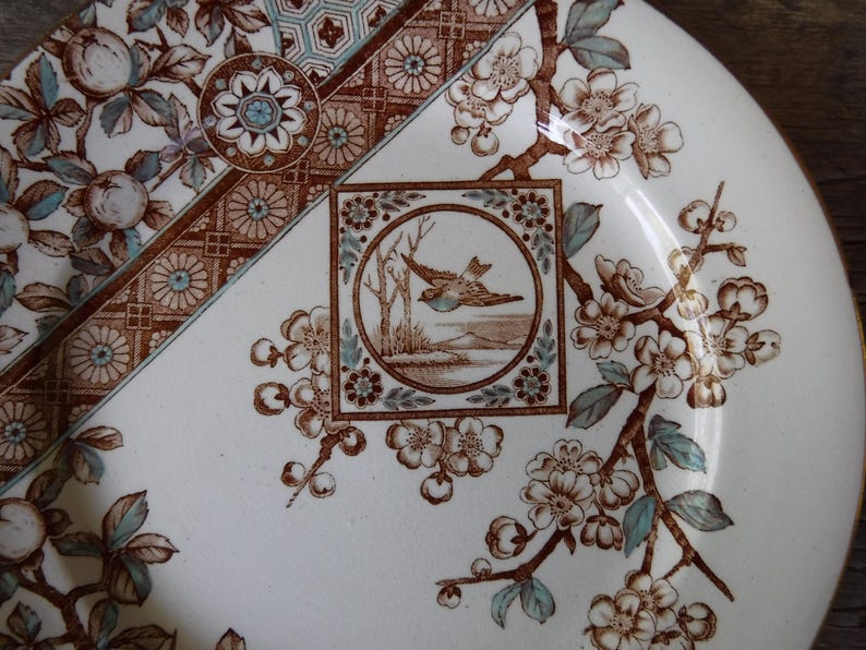 Puede incluir: Un plato de cer&aacute;mica con base color crema y patrones florales en marr&oacute;n y azul claro. Un marco cuadrado central contiene una ilustraci&oacute;n circular de un ave en vuelo sobre un paisaje. El borde presenta motivos geom&eacute;tricos y de frutas.