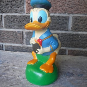 Puede incluir: Una figura vintage de plástico de Donald Duck con un sombrero de marinero azul, una camisa azul y pies naranjas. Está sosteniendo un objeto negro en su mano derecha y parado sobre una base verde.