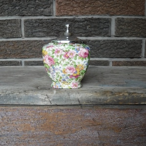 Puede incluir: Un tarro de cerámica decorativo con un estampado floral con rosas rosas, flores amarillas y detalles azules. El tarro tiene una tapa plateada y está sobre una superficie de madera. El fondo es una pared de ladrillo.