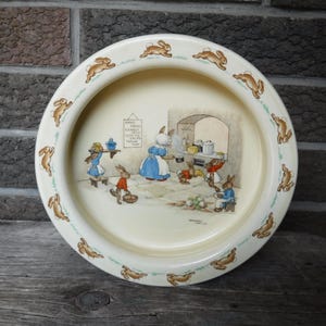 May include: A white ceramic children's bowl with a border of brown bunnies. The center of the bowl features a scene of bunnies in a kitchen with a sign that reads "What We Have To Do Today:  Wash, Peel, Cut, Cook, Serve, Eat, Wash Up, Thank You".