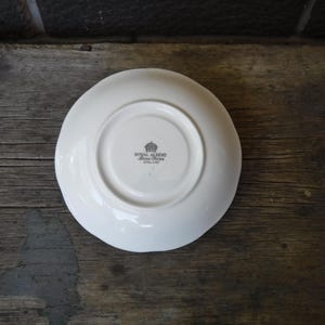 Puede incluir: Un platillo de cer&aacute;mica blanca con una hendidura circular en el centro. La parte inferior presenta un sello negro con un icono de corona y el texto Royal Albert Bone China England. El platillo tiene un acabado liso, brillante y redondo.