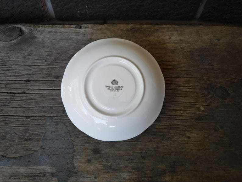 Puede incluir: Un platillo de cer&aacute;mica blanca con una hendidura circular en el centro. La parte inferior presenta un sello negro con un icono de corona y el texto Royal Albert Bone China England. El platillo tiene un acabado liso, brillante y redondo.