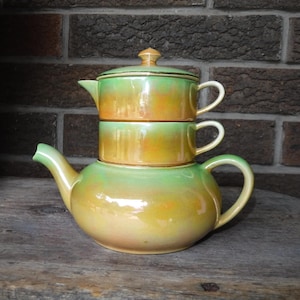 Puede incluir: Una tetera de cerámica de tres niveles con un esmalte verde y naranja. La tetera tiene un cuerpo redondo y una tapa con un pomo en la parte superior. La tetera está sentada sobre una superficie de madera.