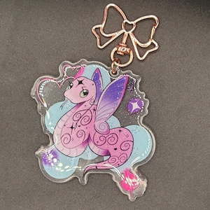 May include: A clear acrylic keyring featuring a pink snake with purple wings and a blue cloud background. The snake has a whimsical design with swirls and a star. The keyring has a rose gold bow-shaped clasp.