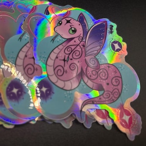 May include: A pile of holographic stickers. Each sticker depicts a pink cartoon snake with butterfly wings, decorative swirls, and a star. The background is light blue, with a rainbow sheen. The stickers are stacked on top of each other.