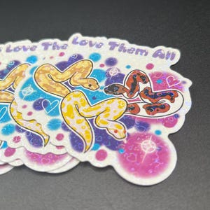 May include: A stack of stickers featuring colorful cartoon snakes with the text "To Love The Love Them All". The stickers have a white border and a holographic finish, with hearts and sparkles.