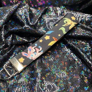 May include: A black wristlet keychain featuring colorful cartoon characters and stars. The wrist strap has a silver metal clasp and a split ring. The background is a shiny, textured black fabric.
