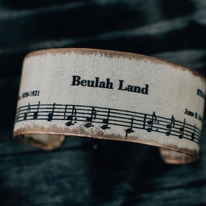 May include: A beige leather cuff bracelet with the words "Beulah Land" printed on it. The bracelet also features a musical staff with notes printed on it.