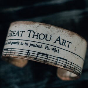 May include: A wide cuff bracelet with a distressed off-white finish featuring black printed text that reads GREAT THOU ART and and greatly to be praised. Ps. 48:1, along with black musical staff lines and notes printed across the surface.